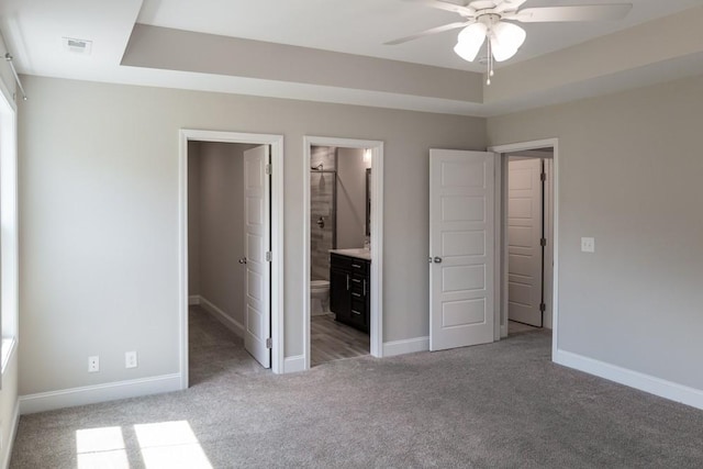 unfurnished bedroom with a raised ceiling, connected bathroom, ceiling fan, and light colored carpet