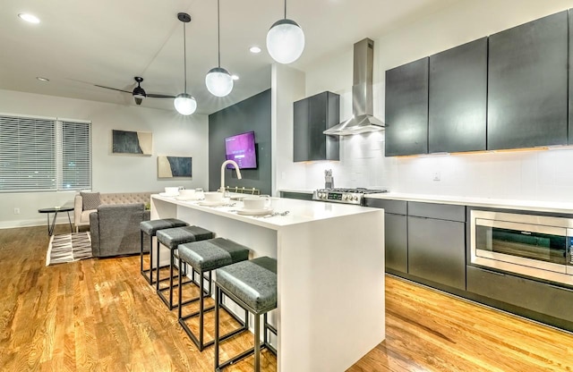 kitchen featuring stainless steel microwave, a center island with sink, wall chimney exhaust hood, ceiling fan, and decorative light fixtures