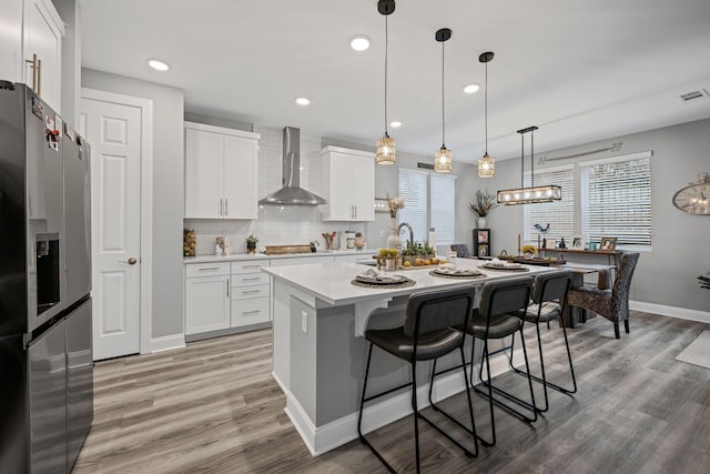 kitchen featuring white cabinets, wall chimney range hood, hanging light fixtures, stainless steel fridge, and an island with sink