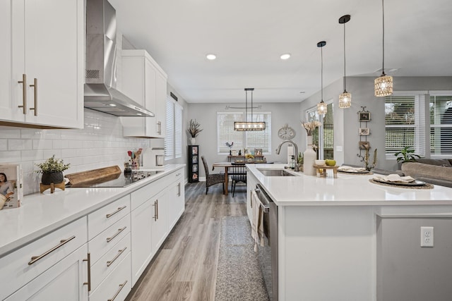 kitchen with black electric stovetop, wall chimney range hood, an island with sink, and hanging light fixtures