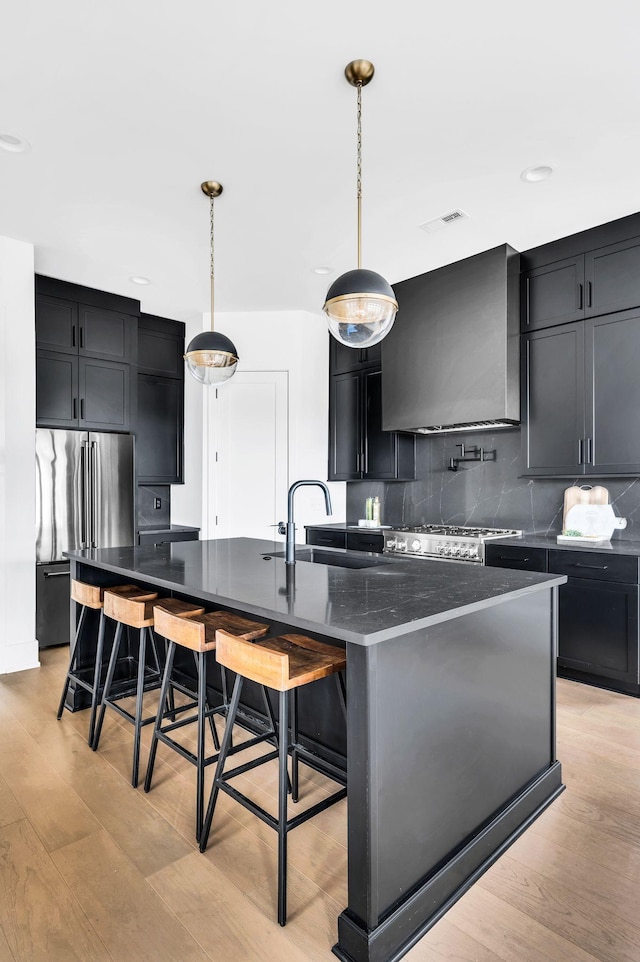 kitchen featuring pendant lighting, wall chimney range hood, an island with sink, high end fridge, and a kitchen bar