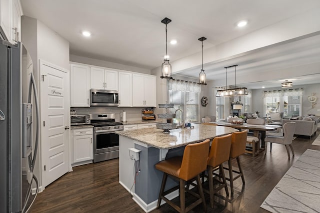 kitchen featuring an island with sink, appliances with stainless steel finishes, decorative light fixtures, light stone counters, and white cabinetry