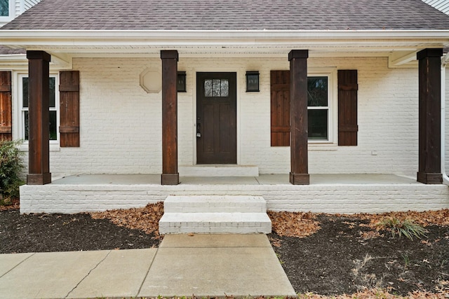 doorway to property featuring covered porch, roof with shingles, and brick siding