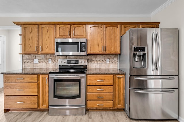 kitchen with decorative backsplash, appliances with stainless steel finishes, ornamental molding, dark stone counters, and light hardwood / wood-style floors