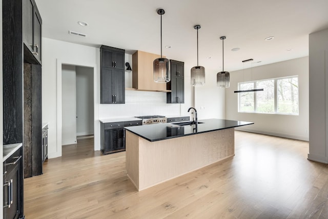 kitchen with wall chimney exhaust hood, sink, range, light hardwood / wood-style floors, and an island with sink