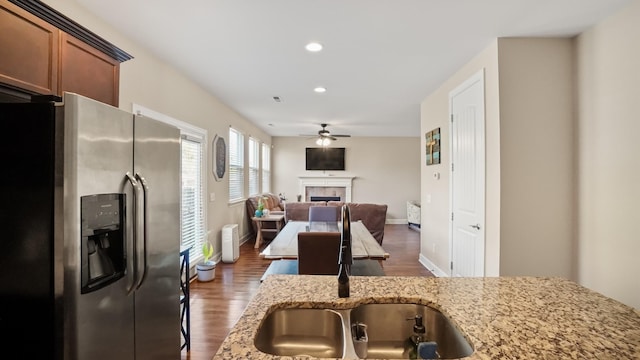 kitchen featuring ceiling fan, sink, light stone counters, stainless steel fridge, and a tiled fireplace