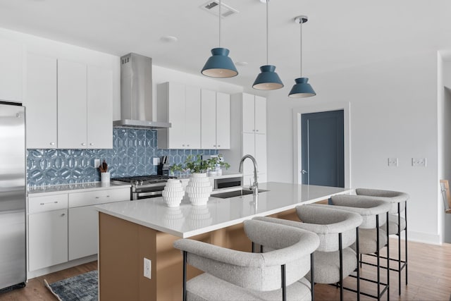 kitchen with white cabinetry, wall chimney range hood, an island with sink, and appliances with stainless steel finishes