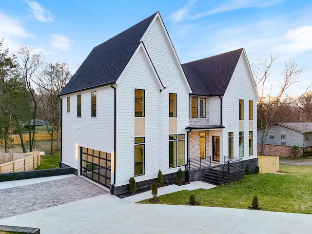 modern farmhouse featuring driveway, stone siding, an attached garage, fence, and a front lawn