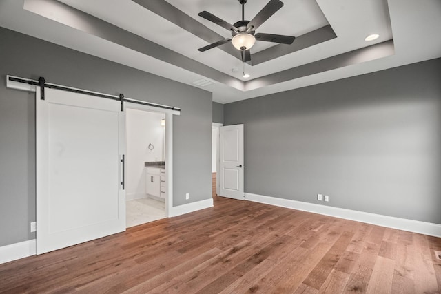 unfurnished bedroom featuring a raised ceiling, ceiling fan, a barn door, connected bathroom, and light hardwood / wood-style floors