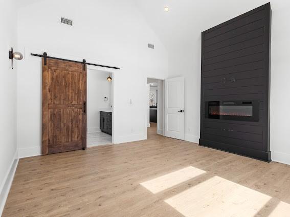 interior space featuring high vaulted ceiling, a barn door, light wood-style flooring, visible vents, and a glass covered fireplace