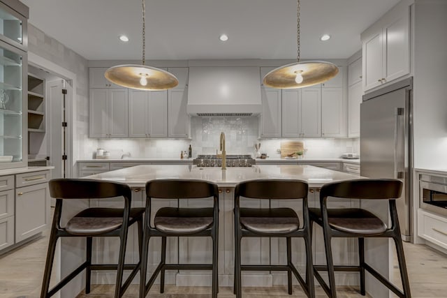 kitchen with a kitchen island with sink, appliances with stainless steel finishes, a kitchen breakfast bar, and premium range hood