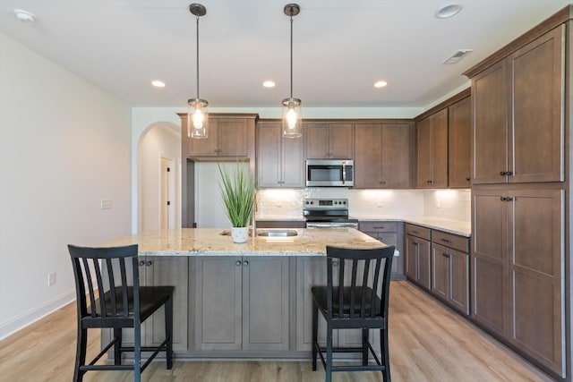 kitchen with light stone countertops, hanging light fixtures, stainless steel appliances, a center island with sink, and light wood-type flooring