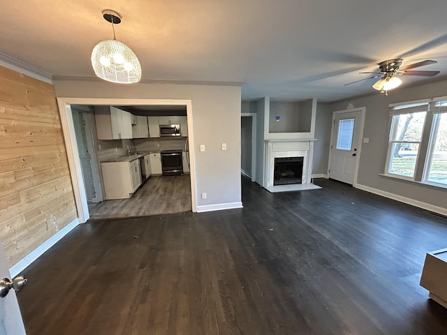 unfurnished living room featuring dark wood-type flooring, a fireplace with flush hearth, wooden walls, baseboards, and ceiling fan with notable chandelier