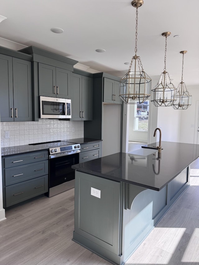 kitchen with light wood-type flooring, a center island with sink, tasteful backsplash, dark countertops, and appliances with stainless steel finishes