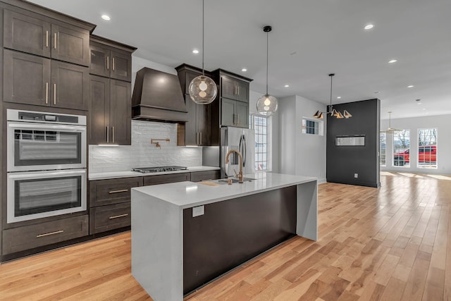 kitchen with stainless steel appliances, premium range hood, a sink, light countertops, and light wood-type flooring