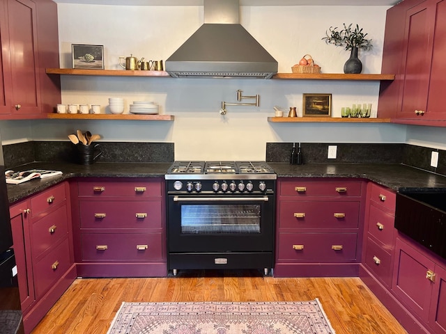 kitchen with light wood-type flooring, range with two ovens, dark stone counters, and wall chimney range hood