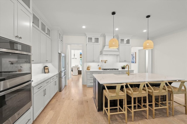 kitchen with white cabinets, custom exhaust hood, stainless steel appliances, an island with sink, and light stone counters