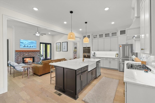 kitchen with a center island with sink, a brick fireplace, appliances with stainless steel finishes, hanging light fixtures, and white cabinets