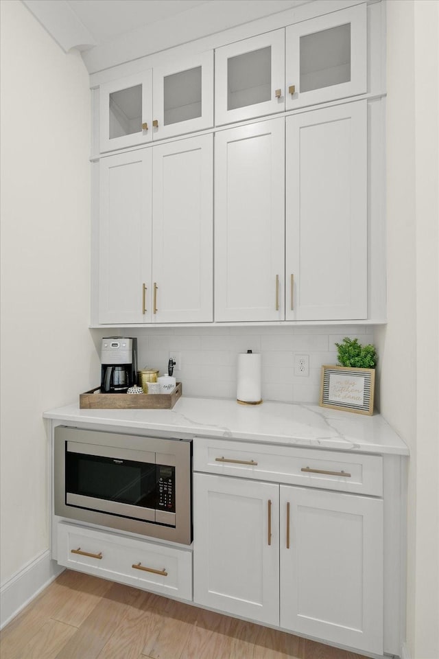 bar featuring stainless steel microwave, tasteful backsplash, white cabinetry, light wood-type flooring, and light stone counters