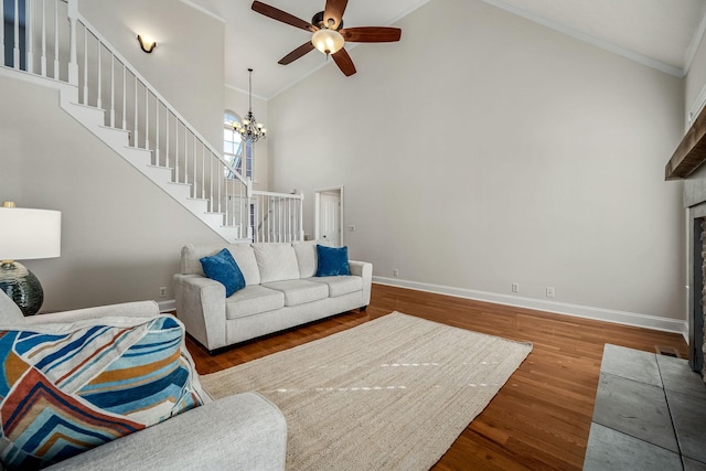 living room with crown molding, high vaulted ceiling, wood-type flooring, a tiled fireplace, and ceiling fan with notable chandelier