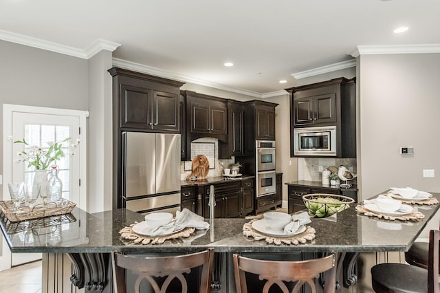 kitchen featuring dark brown cabinetry, stainless steel appliances, tasteful backsplash, dark stone countertops, and a breakfast bar area