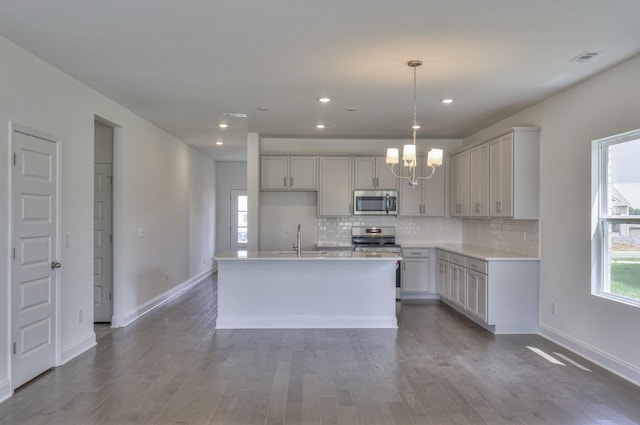 kitchen with gray cabinetry, sink, stainless steel appliances, decorative light fixtures, and a center island with sink