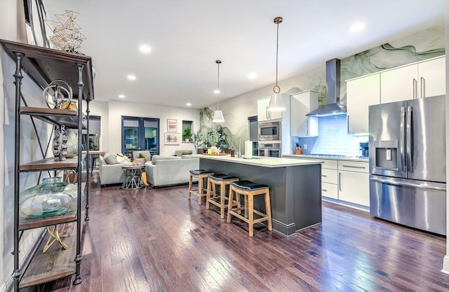 kitchen featuring pendant lighting, appliances with stainless steel finishes, wall chimney exhaust hood, white cabinetry, and a kitchen island with sink