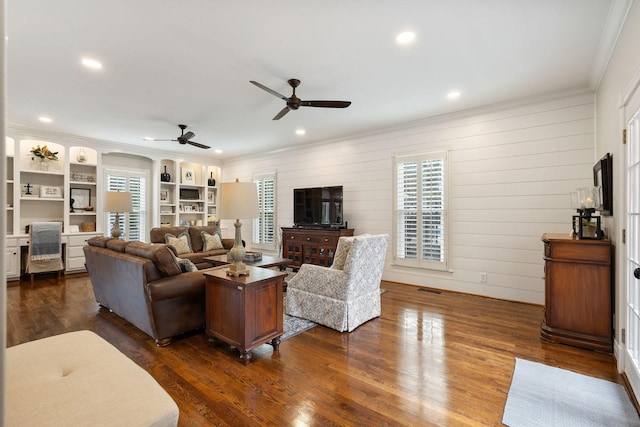 living room with built in shelves, ceiling fan, ornamental molding, and dark wood-type flooring