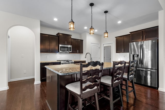 kitchen with light stone counters, appliances with stainless steel finishes, decorative light fixtures, and an island with sink