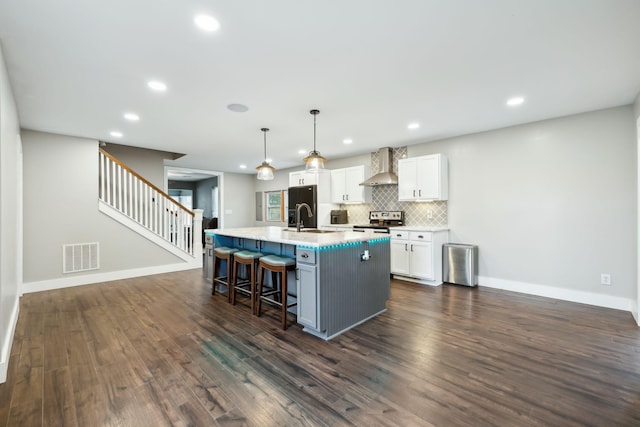 kitchen featuring wall chimney range hood, stainless steel range with electric stovetop, decorative light fixtures, a kitchen island with sink, and white cabinets