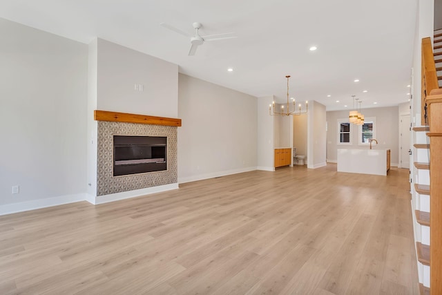 unfurnished living room with baseboards, light wood finished floors, recessed lighting, a tiled fireplace, and ceiling fan with notable chandelier