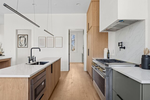 kitchen with sink, appliances with stainless steel finishes, light stone counters, a center island with sink, and wall chimney exhaust hood