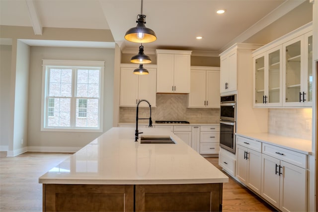 kitchen featuring pendant lighting, a kitchen island with sink, sink, light wood-type flooring, and white cabinetry