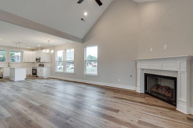 unfurnished living room with a brick fireplace, ceiling fan with notable chandelier, sink, high vaulted ceiling, and light hardwood / wood-style floors