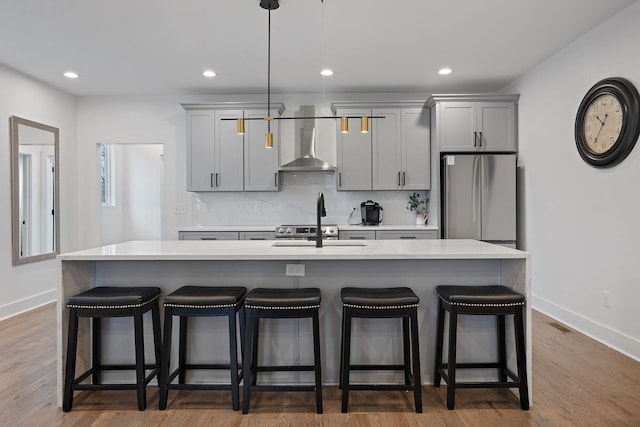 kitchen with gray cabinetry, an island with sink, stainless steel fridge, dark hardwood / wood-style floors, and wall chimney exhaust hood