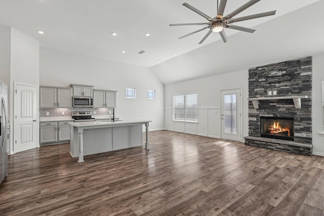 kitchen with gray cabinetry, a kitchen island with sink, a stone fireplace, appliances with stainless steel finishes, and a breakfast bar area