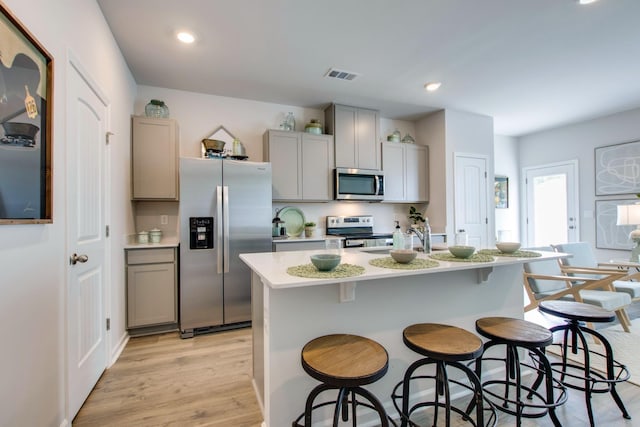 kitchen featuring gray cabinetry, stainless steel appliances, an island with sink, a kitchen bar, and light wood-type flooring