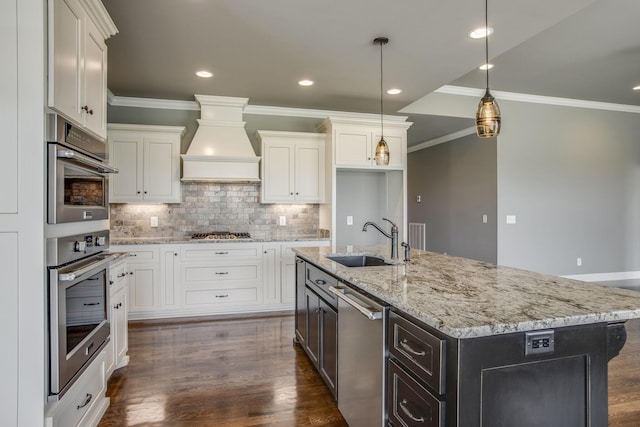 kitchen with sink, appliances with stainless steel finishes, hanging light fixtures, an island with sink, and custom exhaust hood