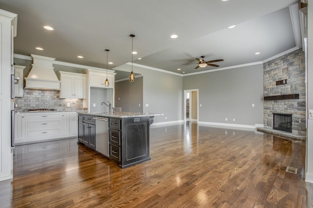 kitchen with custom exhaust hood, a center island with sink, hanging light fixtures, stainless steel dishwasher, and white cabinets