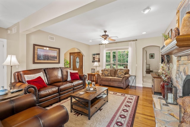 living room with light wood-type flooring, a stone fireplace, and ceiling fan