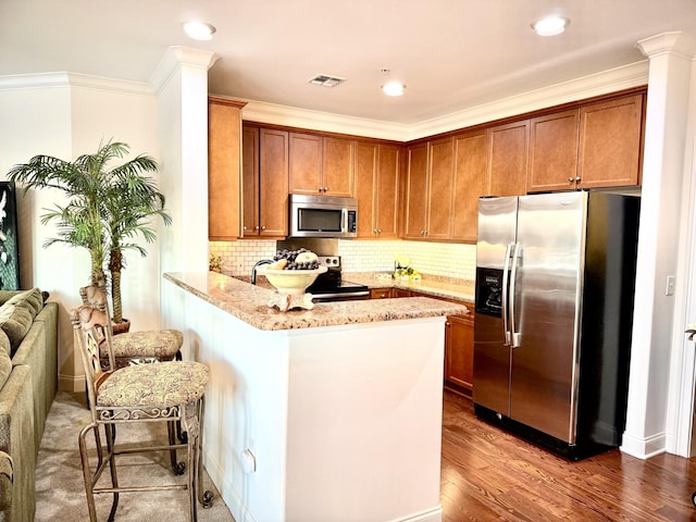 kitchen with kitchen peninsula, light stone countertops, a kitchen bar, appliances with stainless steel finishes, and ornamental molding