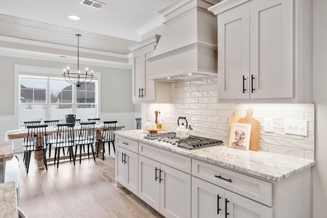 kitchen featuring custom exhaust hood, stainless steel gas cooktop, pendant lighting, a chandelier, and white cabinetry