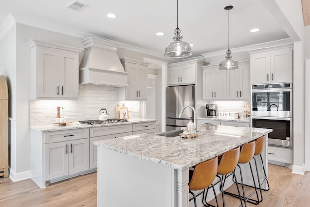 kitchen featuring pendant lighting, a kitchen island with sink, white cabinets, appliances with stainless steel finishes, and custom range hood