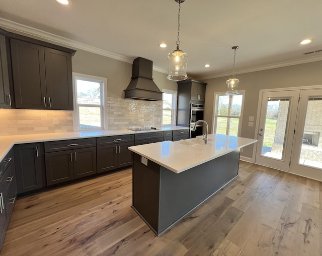 kitchen with double oven, an island with sink, light hardwood / wood-style floors, and custom range hood