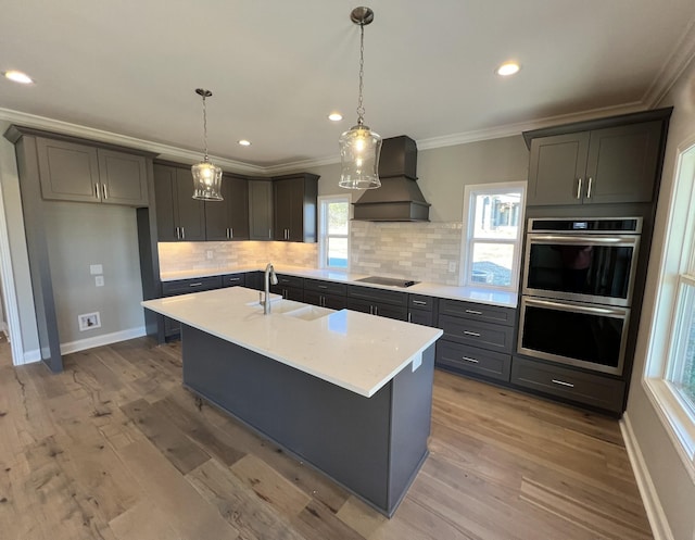 kitchen featuring black electric stovetop, premium range hood, stainless steel double oven, sink, and a center island with sink