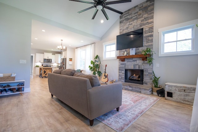 living room featuring ceiling fan with notable chandelier, a stone fireplace, light wood-type flooring, and high vaulted ceiling