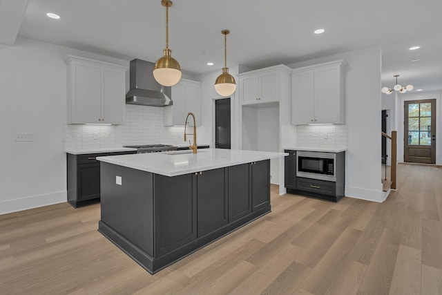 kitchen featuring a kitchen island with sink, built in microwave, wall chimney range hood, decorative light fixtures, and white cabinets