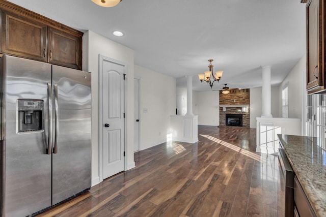 kitchen featuring ornate columns, stainless steel refrigerator with ice dispenser, a fireplace, ceiling fan with notable chandelier, and light stone counters