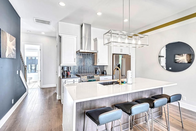 kitchen featuring pendant lighting, high end appliances, a center island with sink, wall chimney range hood, and white cabinetry