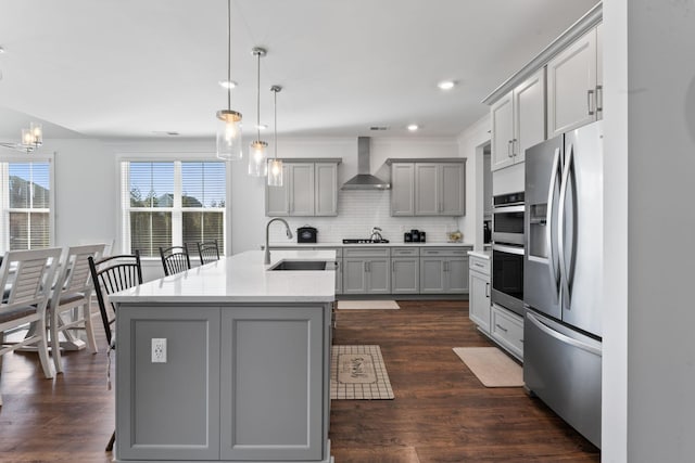 kitchen featuring wall chimney exhaust hood, an island with sink, decorative light fixtures, gray cabinets, and appliances with stainless steel finishes